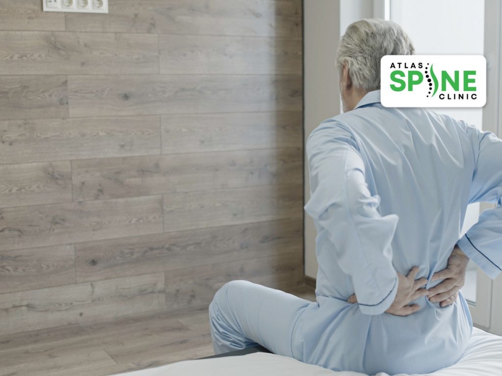 Older man sitting on the edge of a bed holding his lower back, a common sign of spinal stenosis